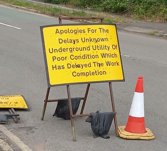 Yellow road sign with confusing apology about delays from unknown underground utility causing work stoppage on the street.