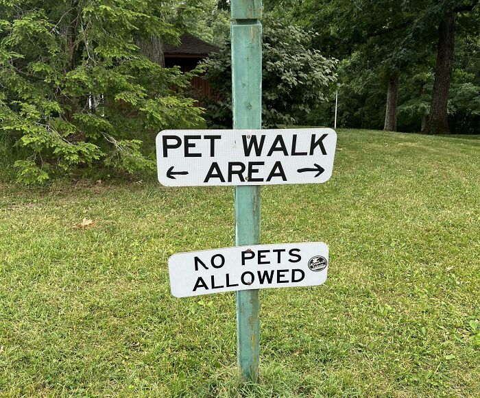 Confusing road signs showing pet walk area arrows alongside no pets allowed sign on a grassy patch.