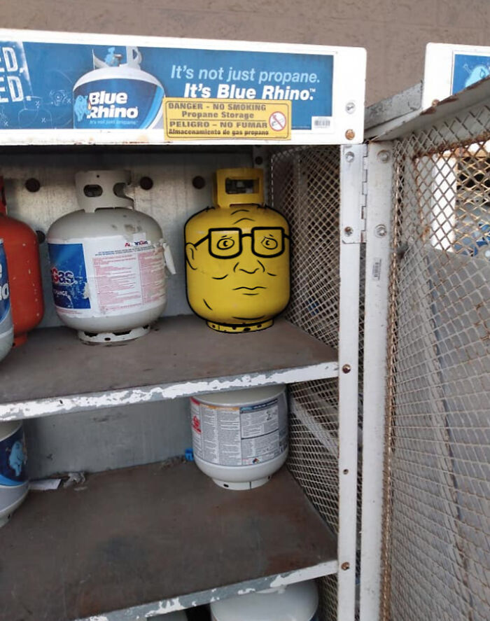 Propane tank painted with a cartoon face, standing out weirdly among regular tanks in a metal storage cage.