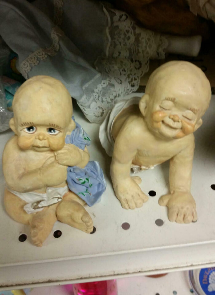Two weird baby dolls with unusual facial expressions sitting on a shelf in a cursed but blessed scene.