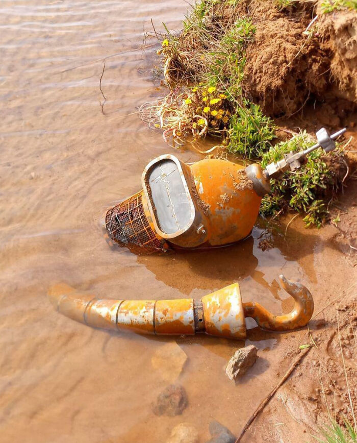 Rusty weird object with hook and gauge half submerged in water near a muddy bank, showing a blessed and cursed vibe.