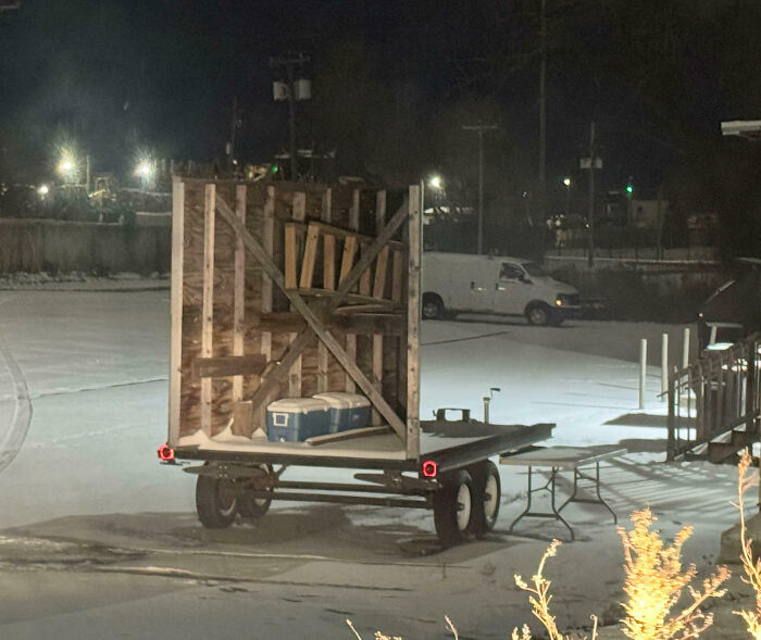 Unphotoshopped pic of a trailer carrying an oddly upright wooden structure and coolers in a snowy parking lot at night