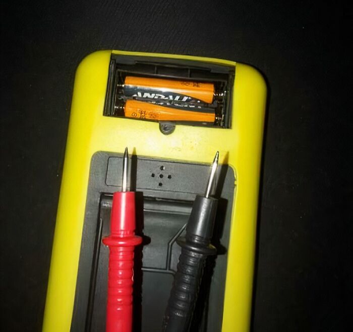 Yellow multimeter with exposed batteries and red and black probes placed incorrectly on the electronic device handling electronics wrong.