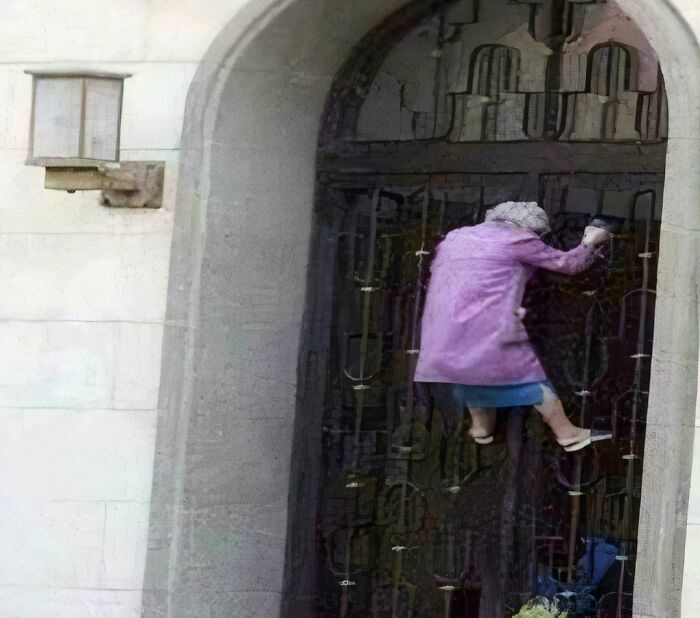 Elderly woman in a purple coat appearing to levitate or climb on a building door, a humorous pleasure of the occult moment.