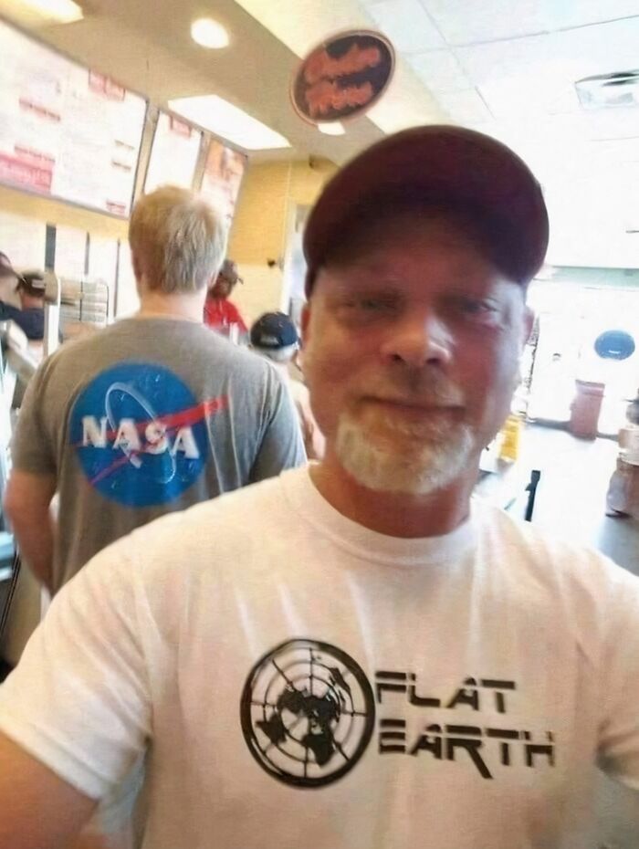 Man wearing a flat earth shirt taking a selfie in a busy fast food restaurant with a NASA shirt in the background.