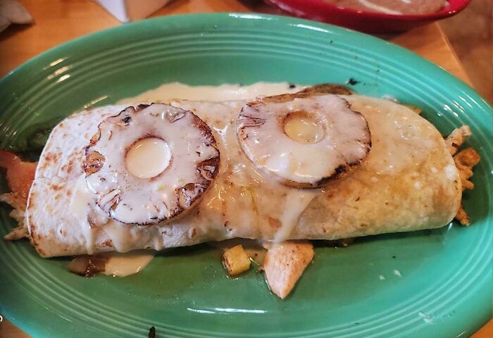 Burrito covered in melted cheese and topped with grilled pineapple slices on a green plate, a cursed and disgusting food picture.