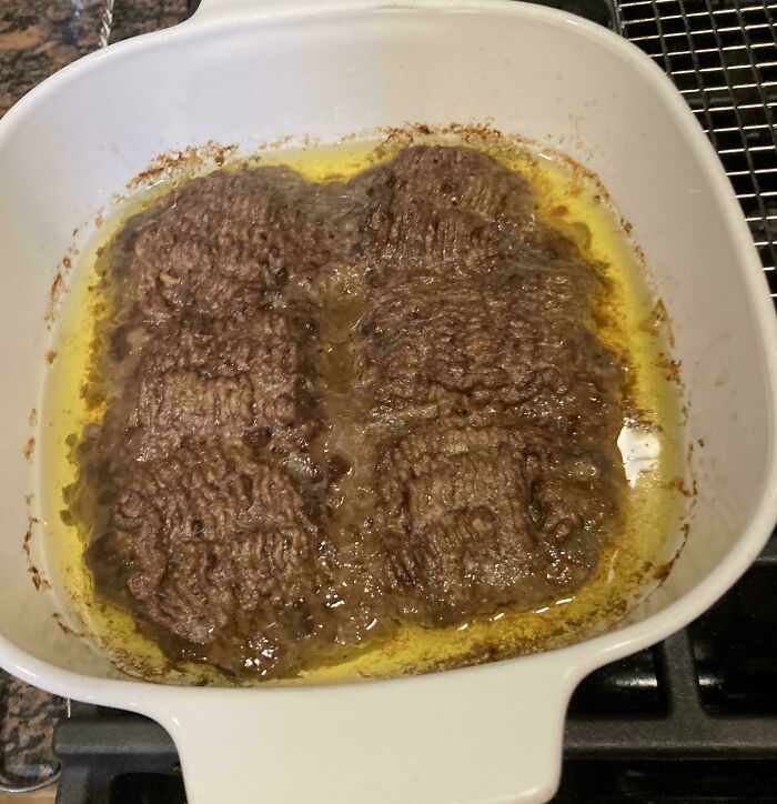 Four overcooked and greasy burger patties sizzling in a white pan filled with oil on a stove.