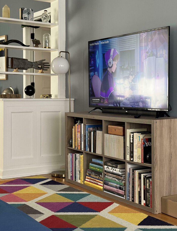 Black and white cat seamlessly merged into the background on a shelf below a TV in a colorful living room.