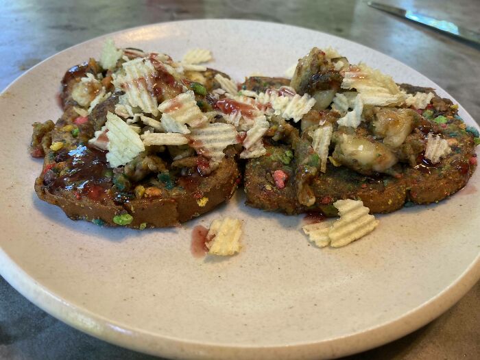 Close-up of cursed and disgusting food featuring soggy toast topped with chips, nuts, and an unappetizing sauce on a beige plate.