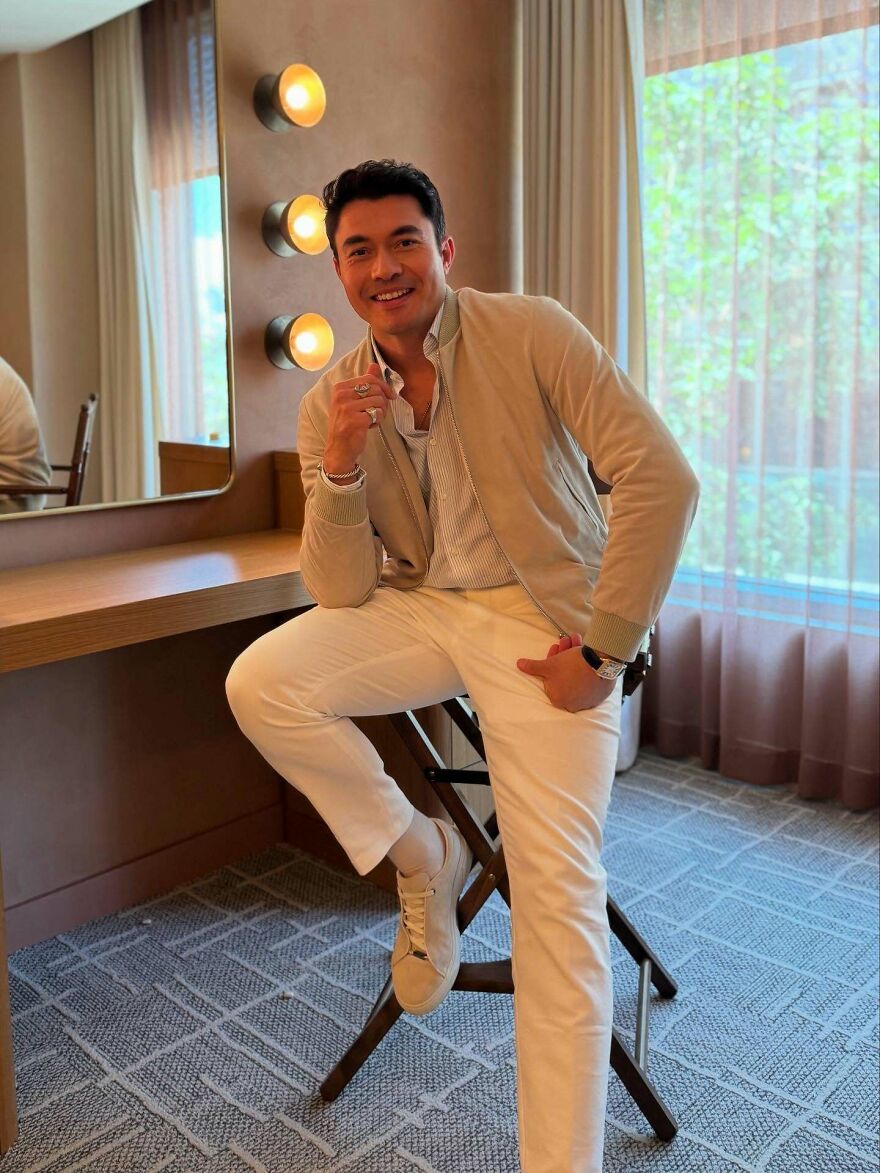 Henry Golding sitting on chair in beige jacket and white pants, smiling confidently in a well-lit room, handsome man in the world.