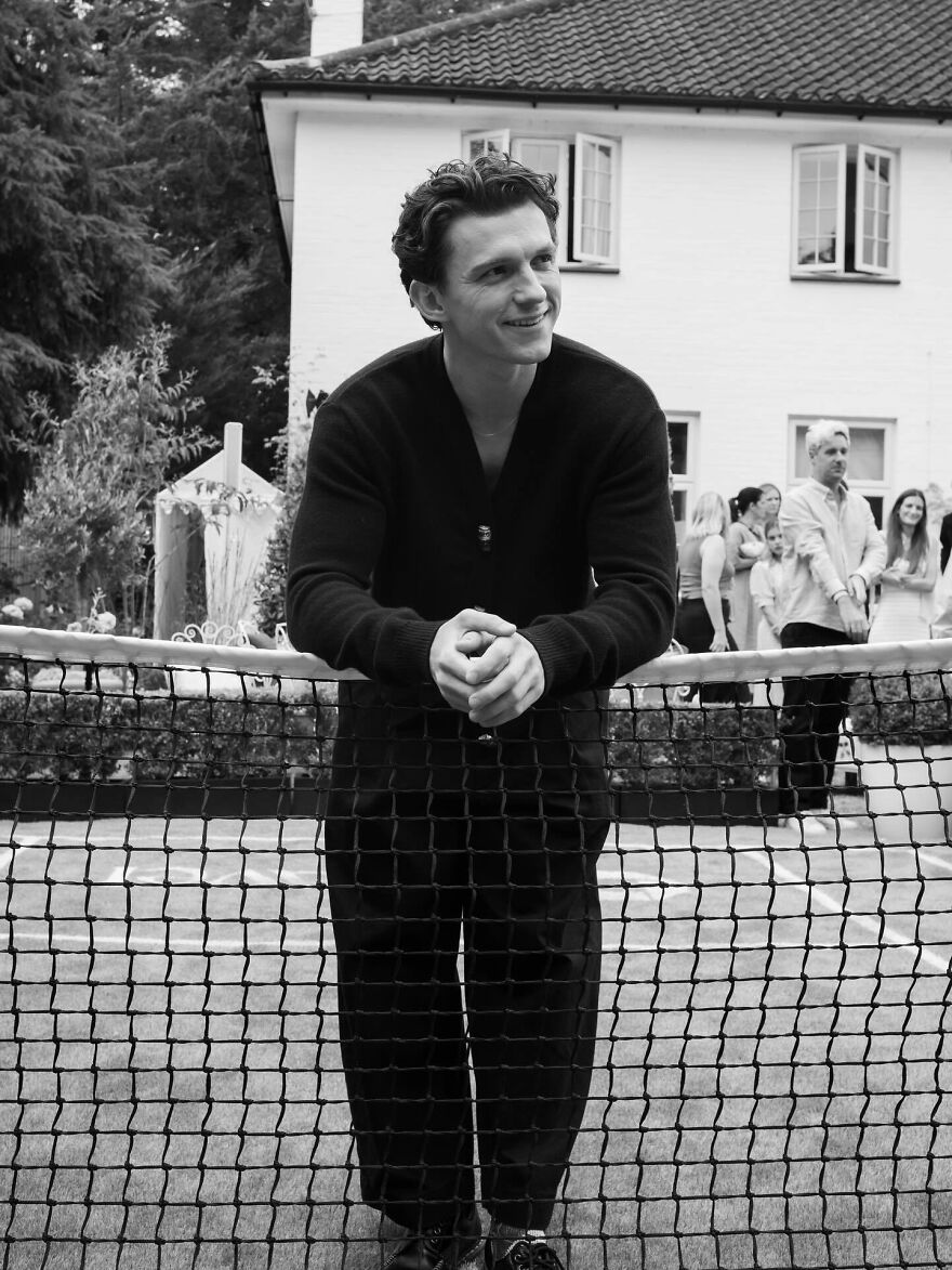 Young Tom Holland smiling and leaning on a tennis net outdoors, representing the most handsome man in the world 2025.