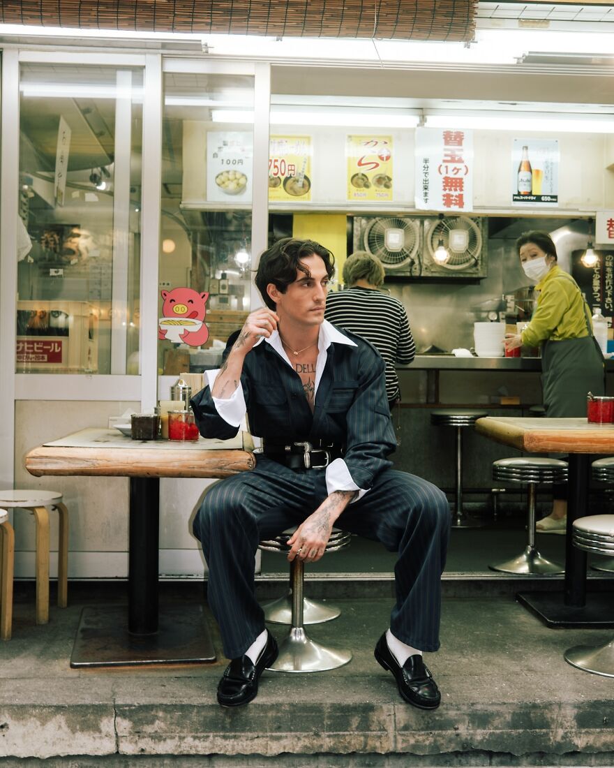 Damiano David man seated outside casual eatery, wearing a black suit and white shirt, embodying the most handsome man in the world vibe.