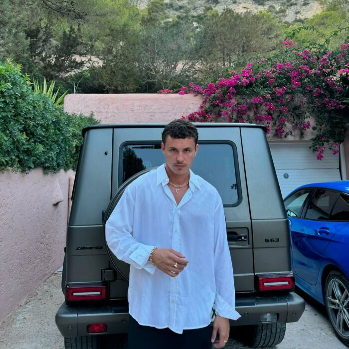 Young man wearing a white shirt posing confidently in front of a luxury SUV, representing most handsome man in the world 2025.