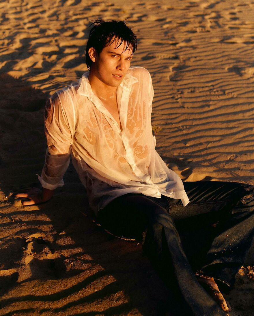 Young Nicholas Galitzine wearing a wet white shirt sitting on sand at sunset, representing the most handsome man in the world 2025.