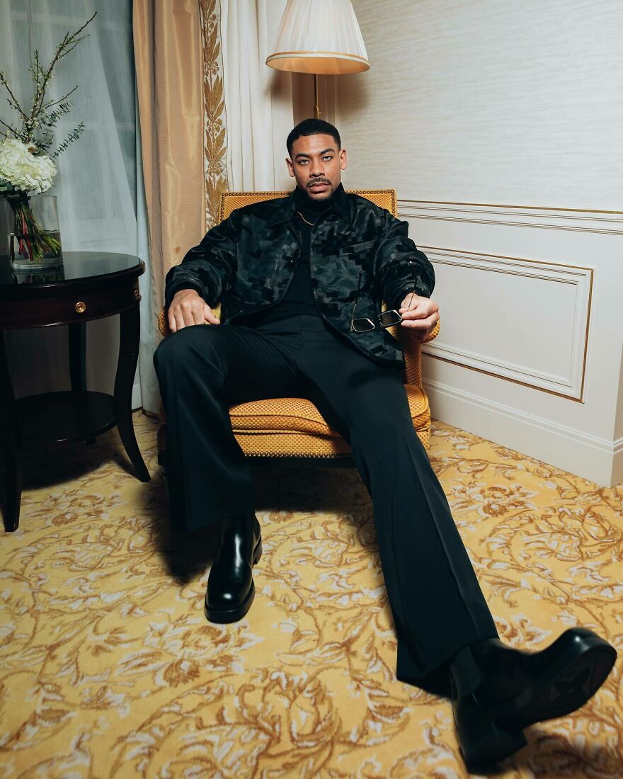 Aaron Pierre sitting confidently in black outfit holding sunglasses indoors, representing most handsome man in the world 2025.