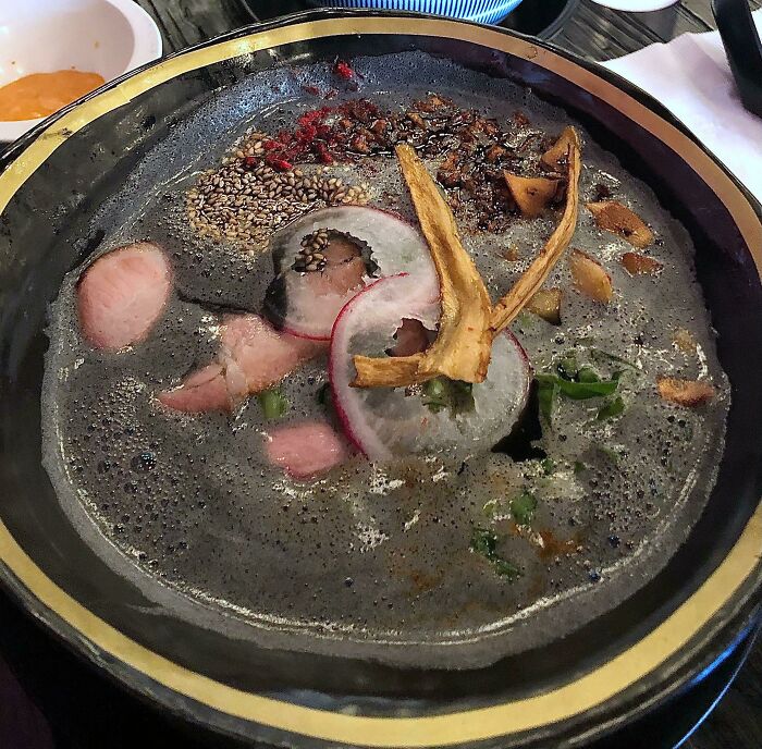 Black, murky soup with radish slices, sesame seeds, and garnishes in a bowl, a cursed and disgusting food picture.