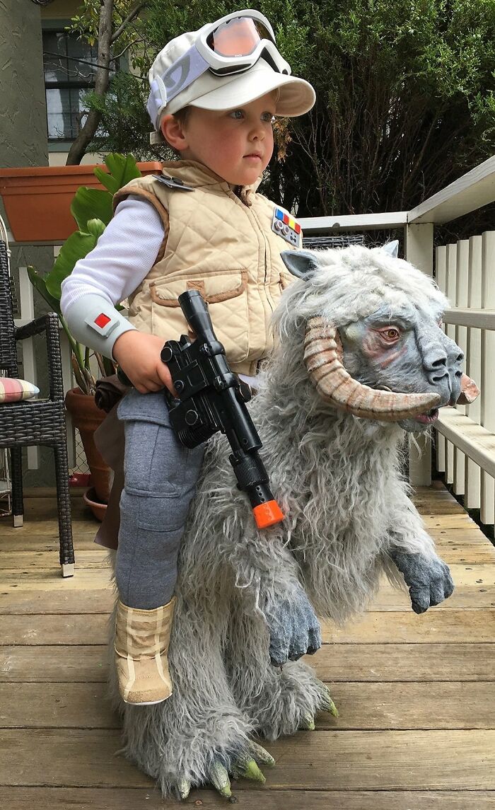 Boy in a creative kids' Halloween costume riding a detailed furry creature with horns and holding a toy gun outdoors.