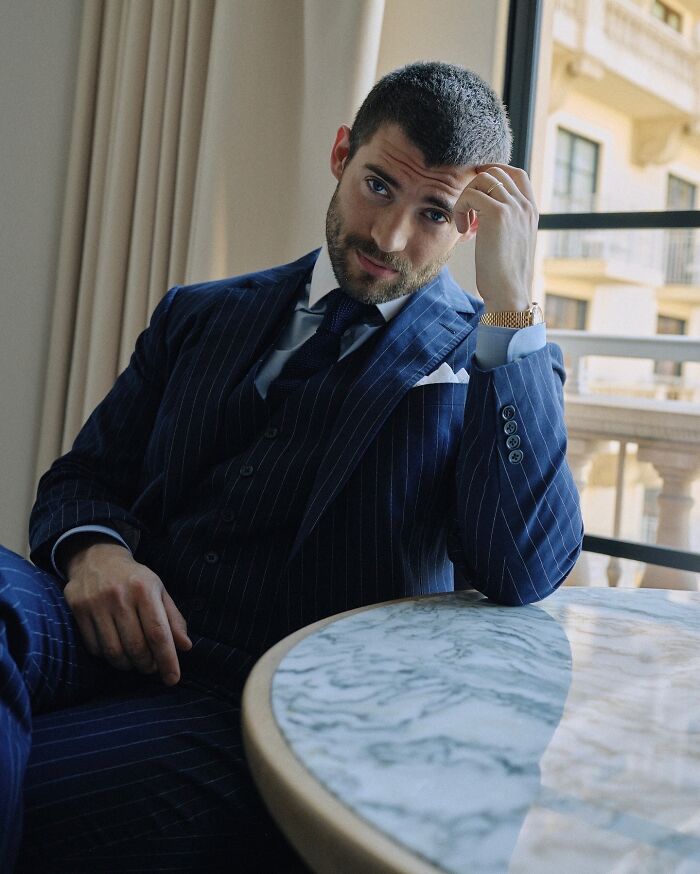 Man in a stylish pinstripe suit sitting by a marble table, embodying the most handsome man in the world 2025 look.