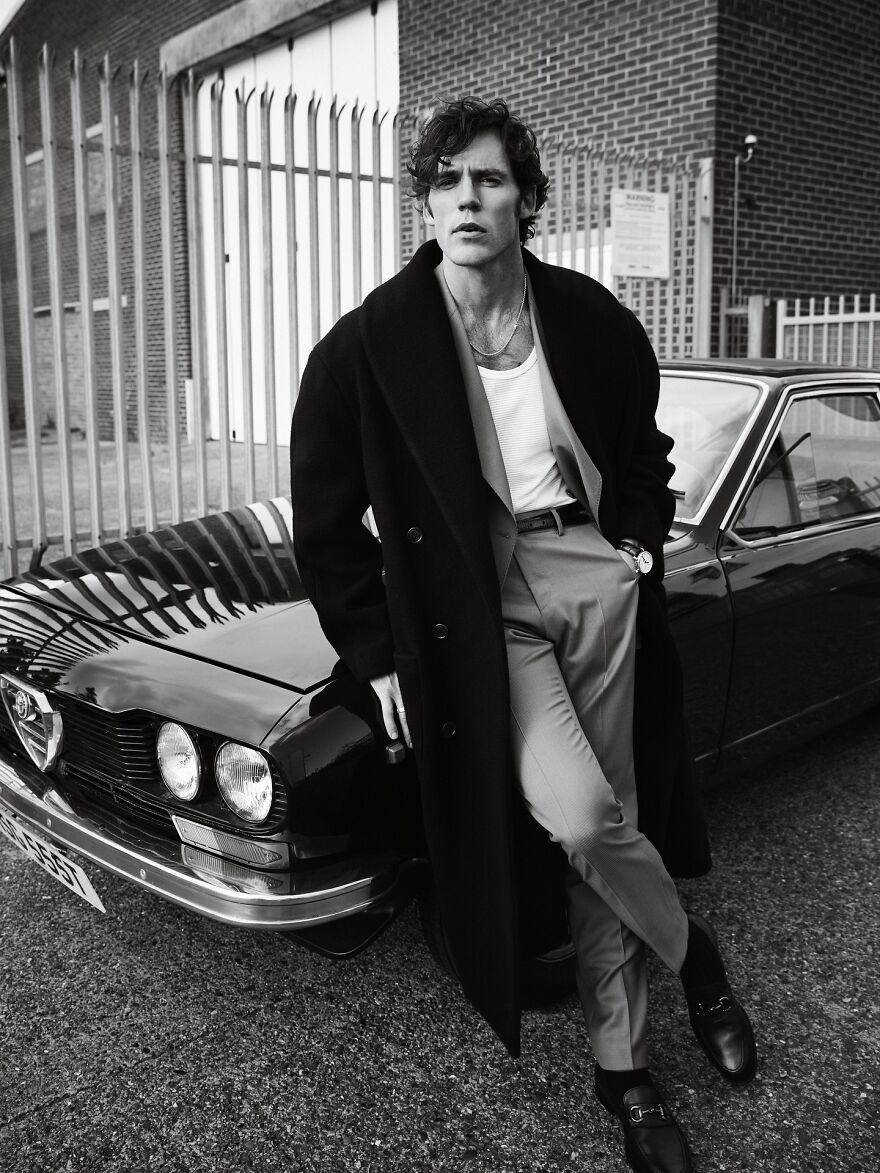 Sam Claflin in a long coat leaning on a vintage car, embodying classic charm and handsome man fashion 2025.