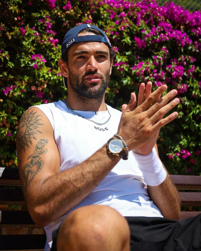Man with tattoos and beard wearing a cap and watch sitting outdoors with flowers in the background, representing most handsome man.