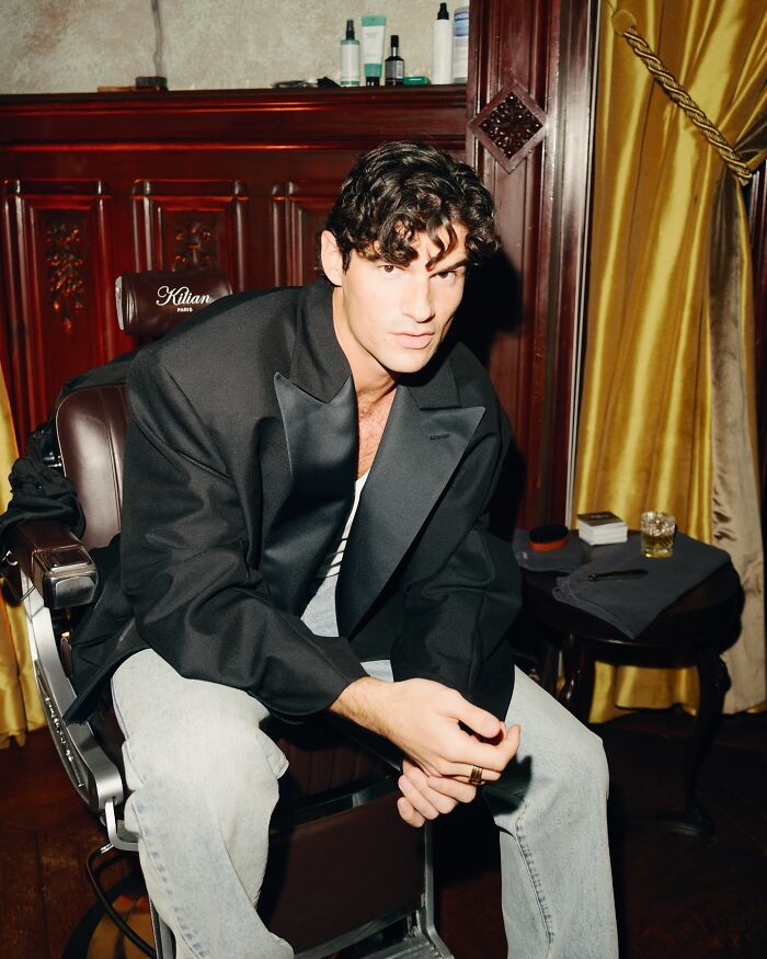 Young handsome man with curly hair wearing a black blazer, sitting in a vintage chair in a stylish indoor setting.