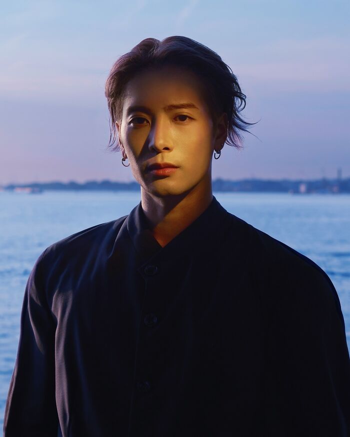 Young man by the water at sunset, wearing dark clothing and earrings, embodying the most handsome man in the world 2025.