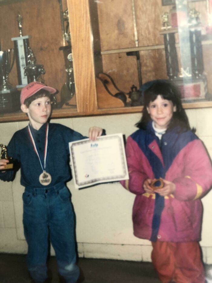 Two kids in front of a trophy case holding a certificate and a medal in a family photo that’s cringed at online.