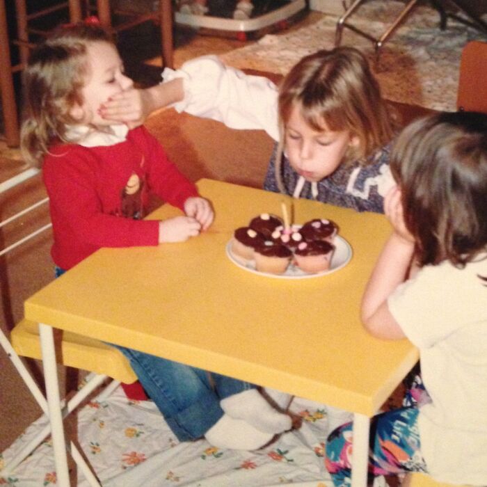 Niños en una foto familiar incómoda celebrando con pastelillos en una mesa amarilla durante una reunión familiar.