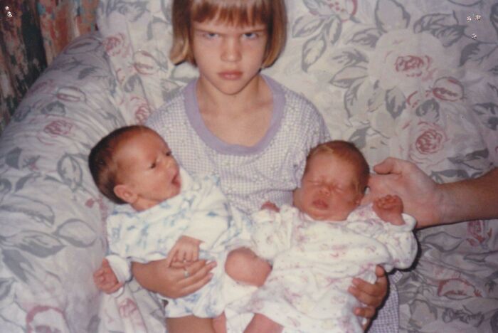 Niña con expresión seria sosteniendo dos bebés en un sofá con funda estampada en una foto familiar incómoda.
