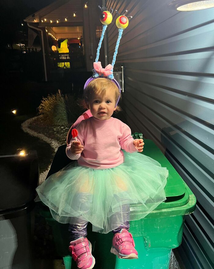 Toddler in an adorable kids Halloween costume with eye stalks, pink sweater, green tutu, and pink shoes sitting outside at night.