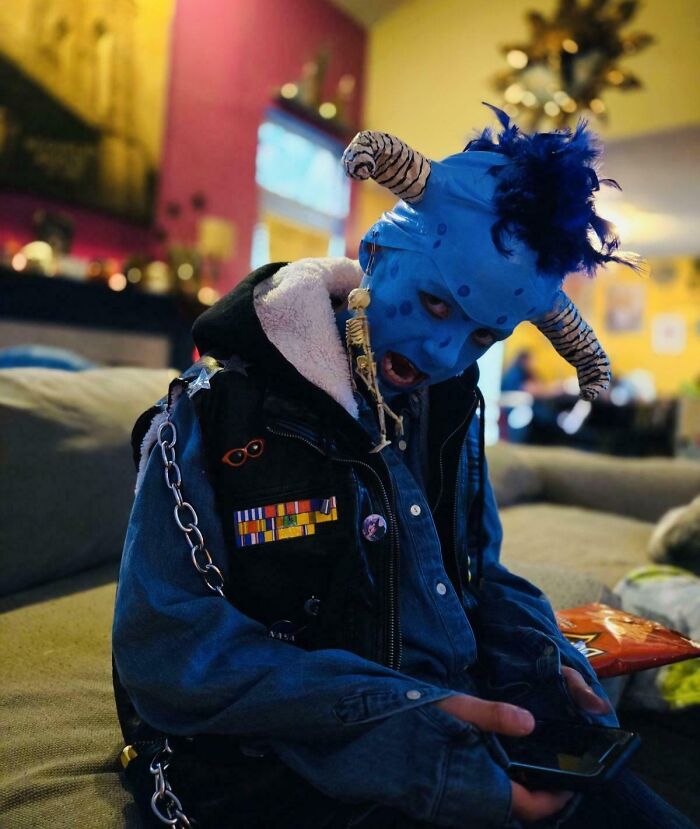 Child in a creative blue demon costume sitting on a couch with Halloween decorations in the background, kids Halloween costume ideas.
