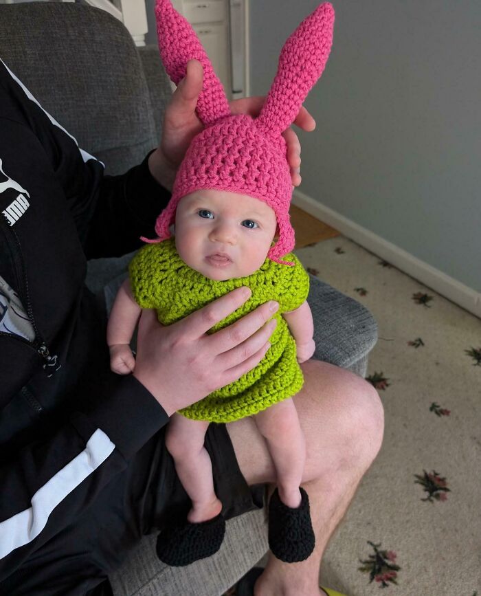 Baby dressed in a handmade crochet kids Halloween costume with pink bunny ears and a green outfit sitting on an adult's lap.