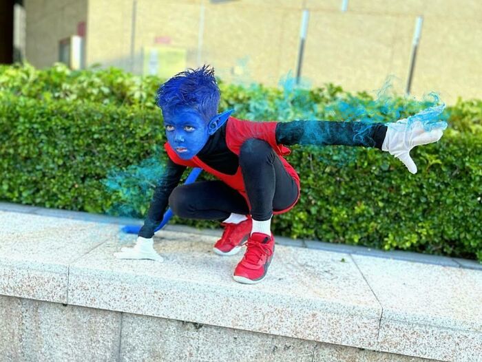 Child in a creative kids Halloween costume with blue face paint and red outfit posing outdoors on a stone ledge.