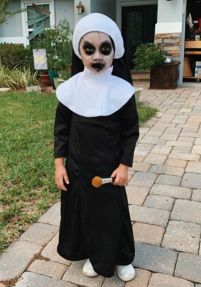 Child in spooky kids Halloween costume wearing black and white nun outfit with dark face paint outdoors on a paved walkway.