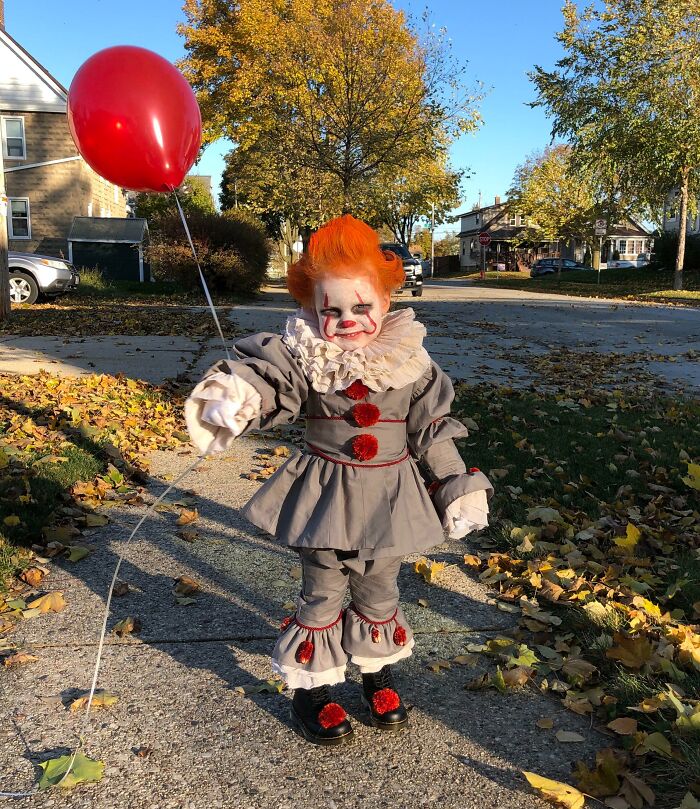 Child in a creative kids Halloween costume holding a red balloon outdoors on a fall day with autumn leaves.
