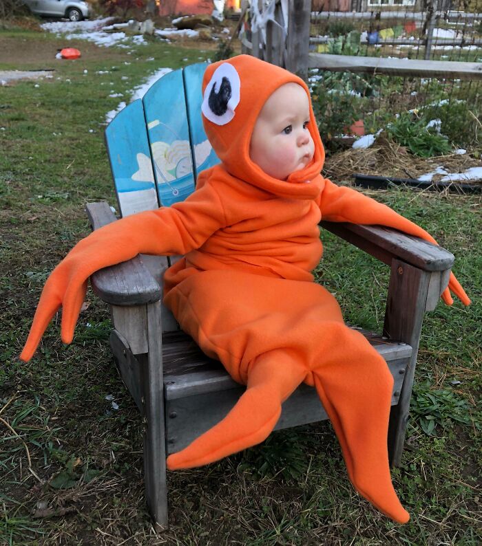 Toddler sitting in a wooden chair wearing an orange Halloween costume resembling a cartoonish crab or lobster.