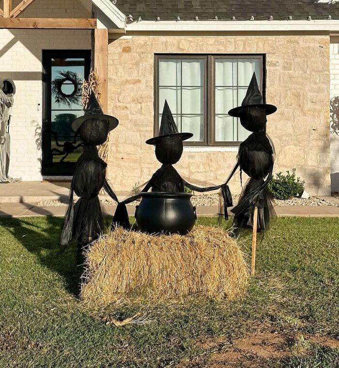 Three black witch decorations holding hands around a cauldron on a hay bale in outdoor Halloween decorations.