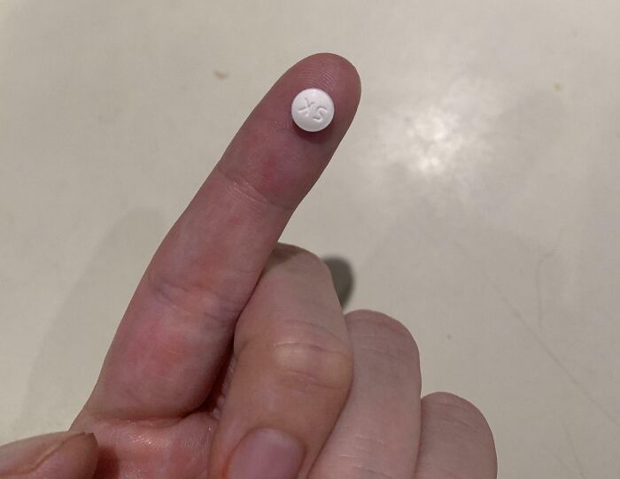 Small white pill marked XS balanced on the fingertip, illustrating harsh realities of American healthcare medication access.
