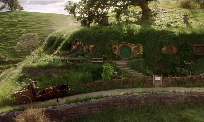 Hobbit hole in a lush green imaginary world with a horse-drawn cart traveling on a curved stone path.