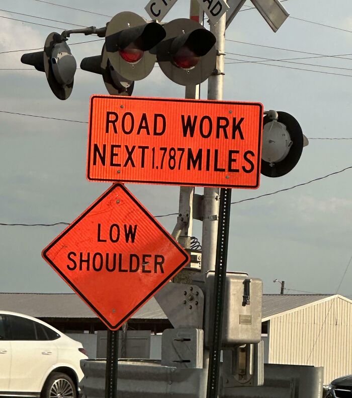 Confusing road signs warning of road work for 1.787 miles and low shoulder near a railroad crossing.