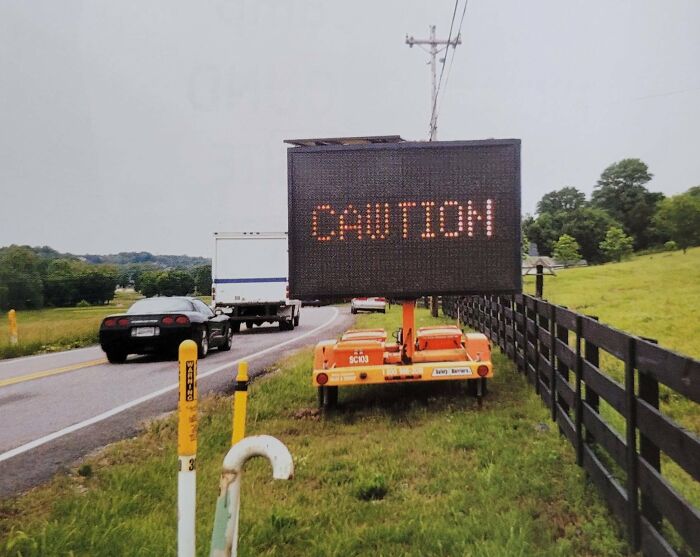Electronic road sign displaying caution on a rural road with cars and a fence, illustrating confusing road signs.