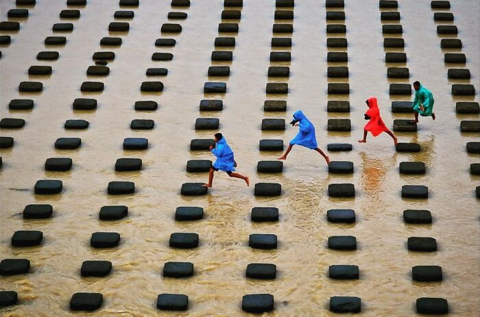 Children wearing colorful raincoats jumping across stepping stones in water, captured through Harjanto Sumawan’s candid photos.
