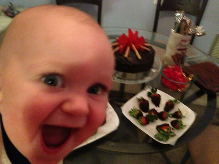 Close-up of a baby photobombing a dessert table with chocolate cake and strawberries, creating a funny photobomb moment.