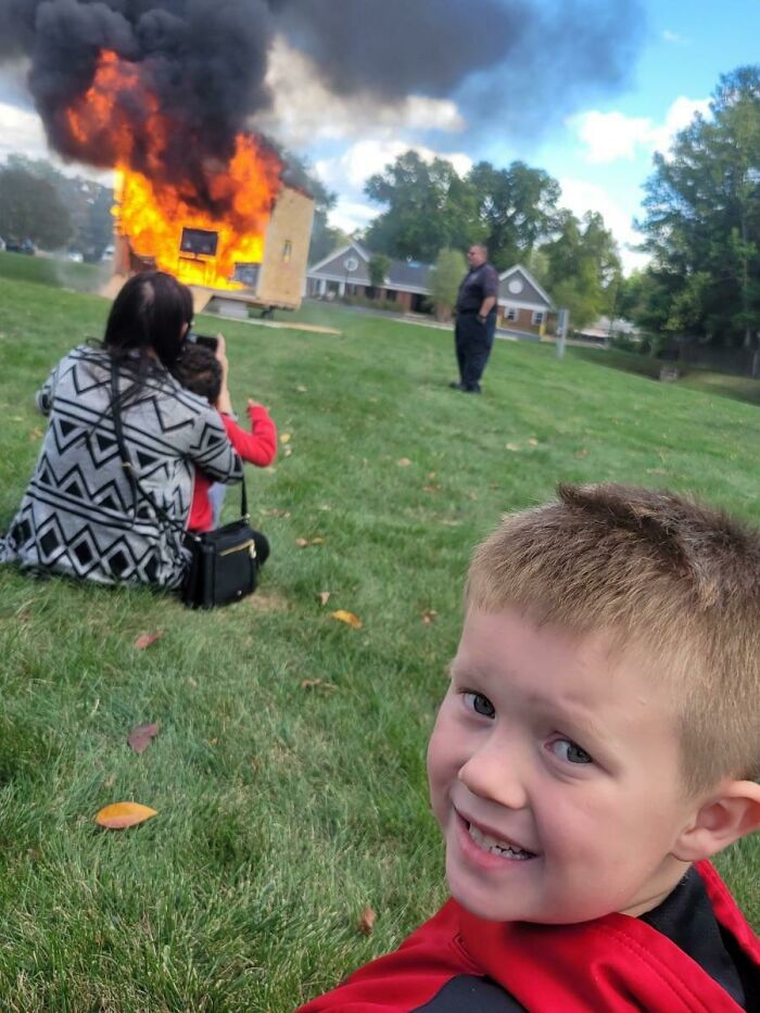 Child smiling in a photobomb near a burning structure with smoke as a dramatic background in an outdoor setting.