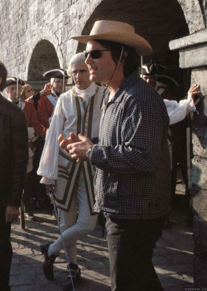 Man wearing sunglasses and a cowboy hat photobombs historic reenactors dressed in colonial costumes outdoors.