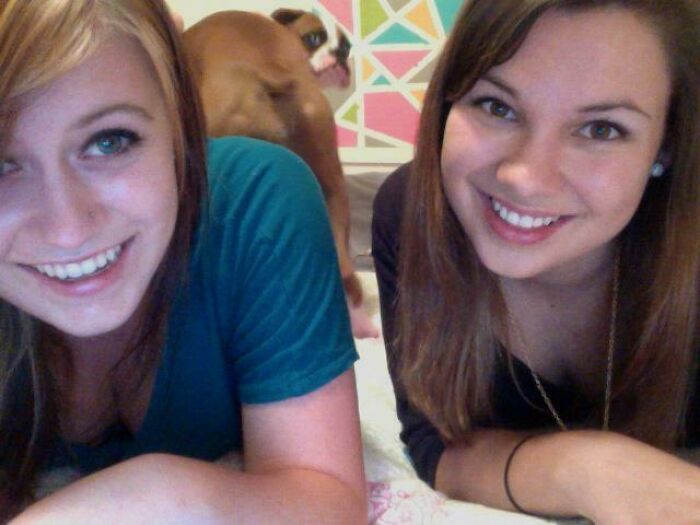 Two smiling women lying down with a photobombing dog making a funny face in the background.