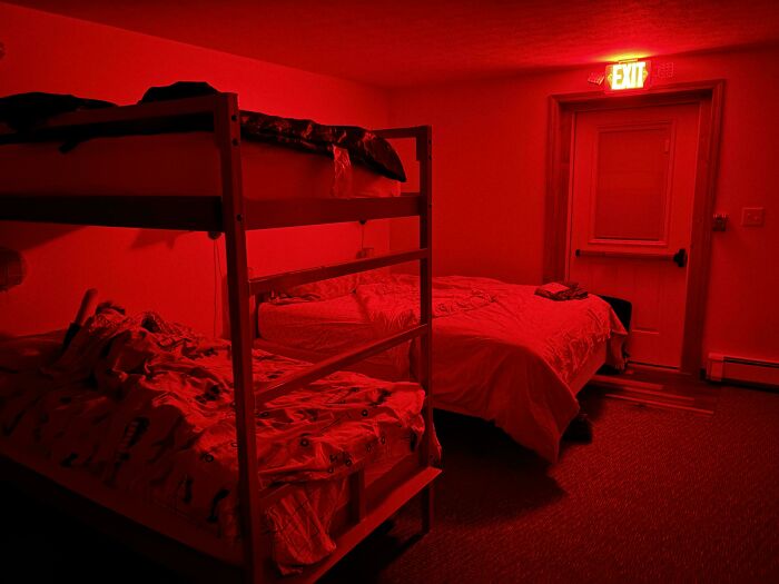 Bunk beds and a single bed in a dimly lit red room with an illuminated exit sign above the door, testing patience.