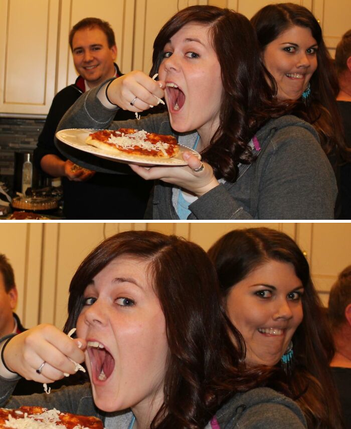 Woman eating pizza while another person photobombs with a funny face in a candid kitchen moment.