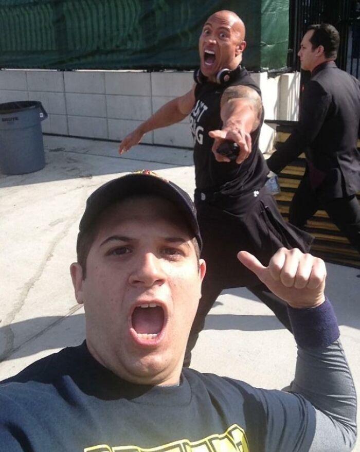 Man taking selfie with a loud photobomb from a muscular man making a shocked face and pointing at the camera outdoors.