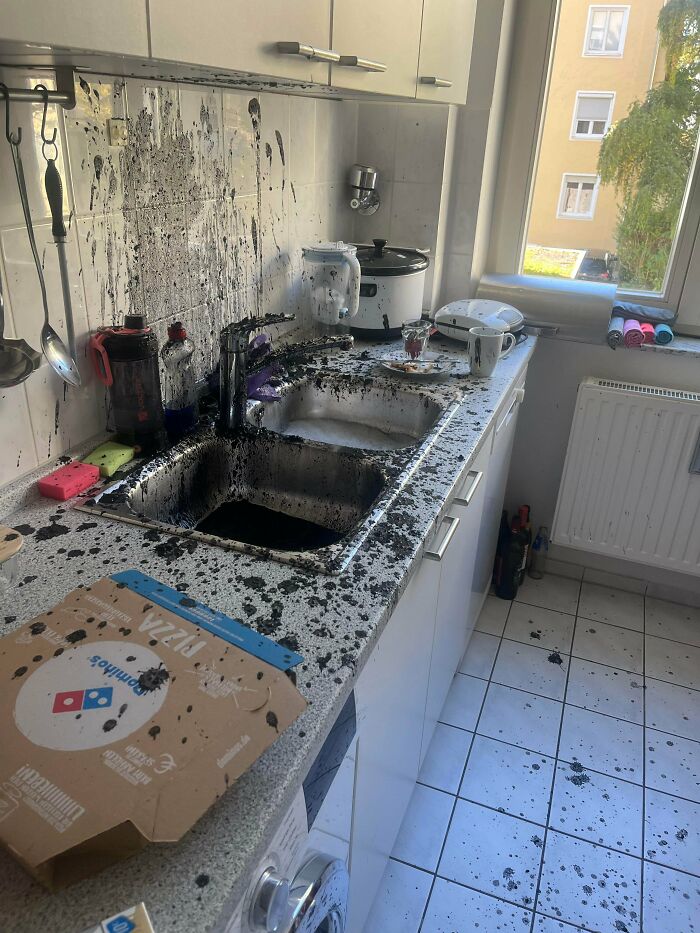 Kitchen sink and countertop covered in spilled black liquid stains, a pizza box nearby, showing mildly infuriating mess testing patience.